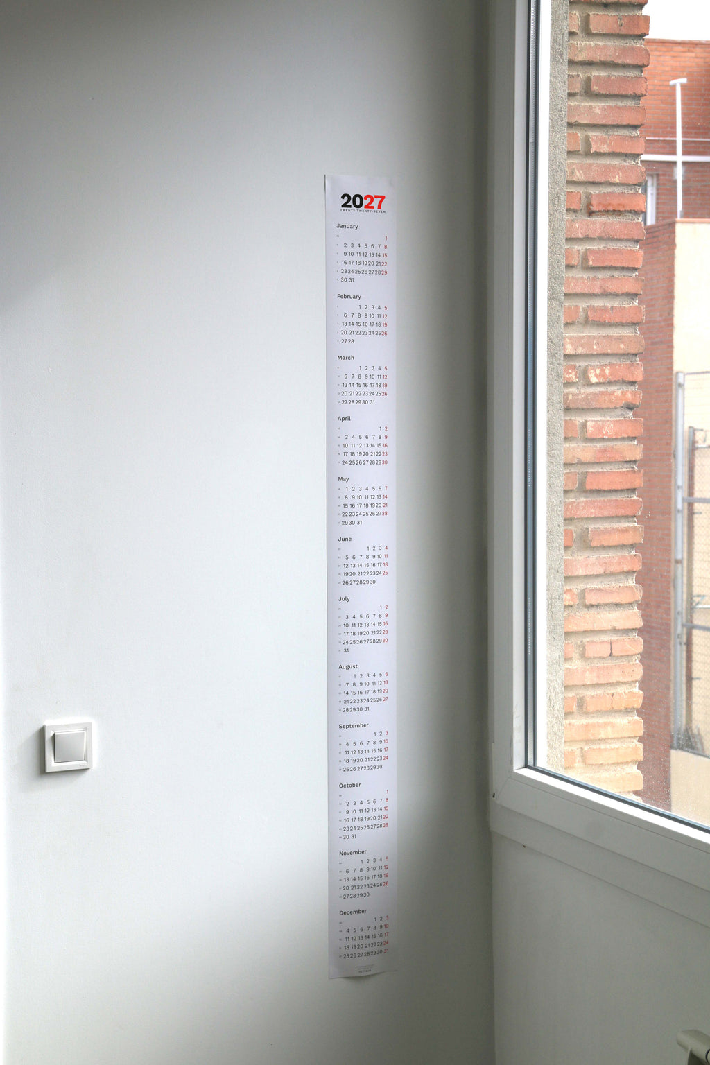 2027 Vertical Calendar by Octàgon Design. The calendar hanging on a wall beside a window.