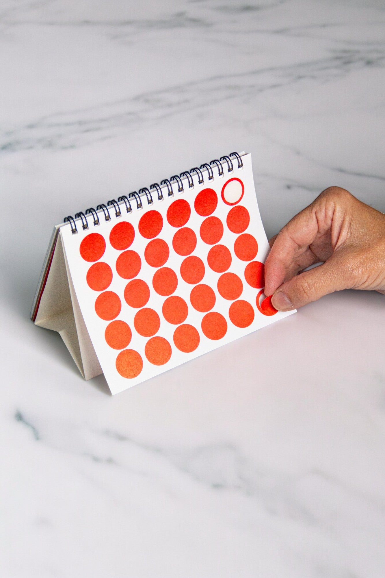 2027 Vinçon desk calendar by Octàgon Design. A hand putting a round red sticker to mark a date.