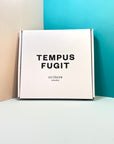 Box with "Tempus fugit" print leaning on a colored wall | Caja estampada "Tempus fugit" apoyada en una pared de colores. | Caixa estampada "Tempus fugit" recolzada en una paret de colors.