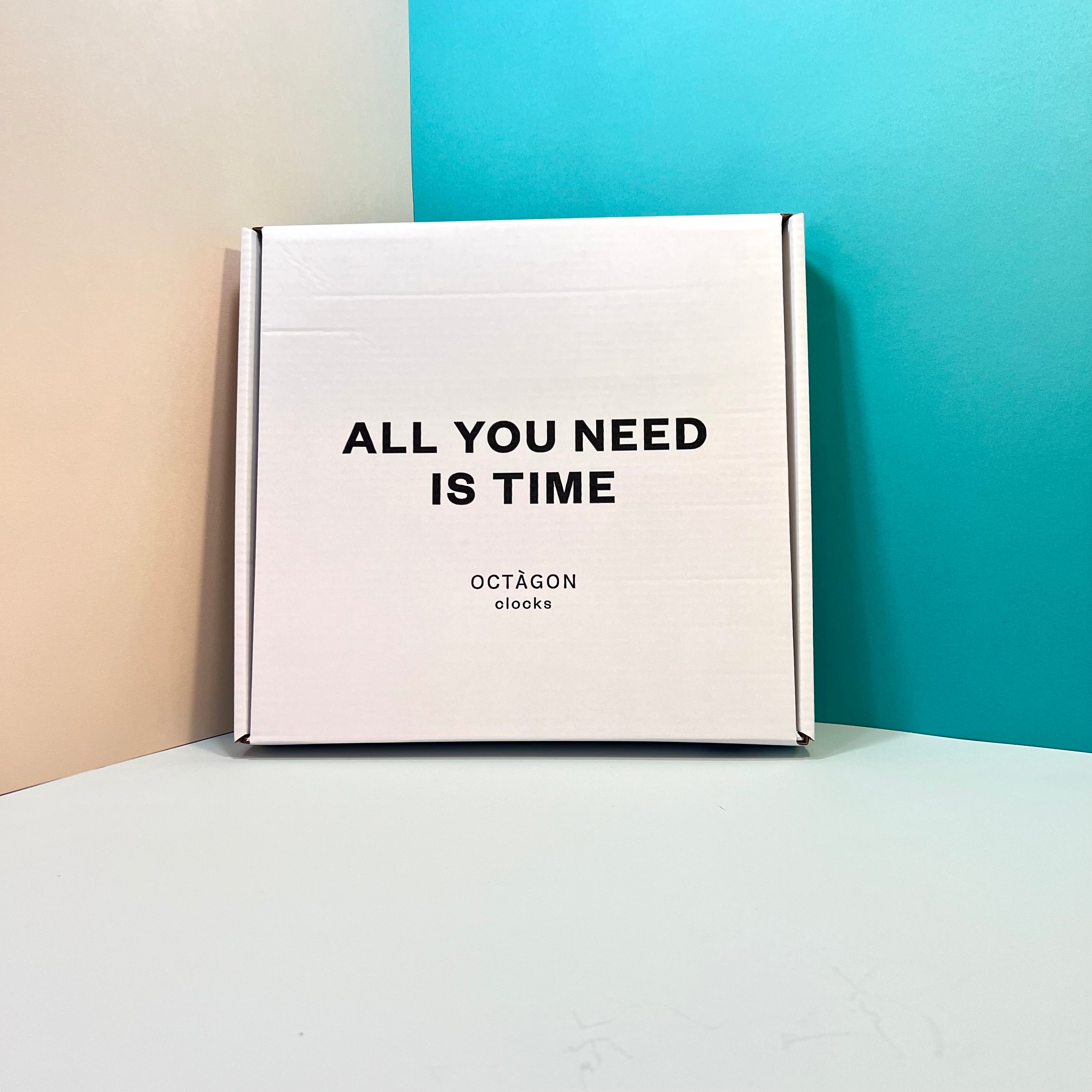 Box with "All you need is time" print leaning on a colored wall | Caja estampada "All ypu need is time" apoyada en una pared de colores. | Caixa estampada "All you need is time" recolzada en una paret de colors.