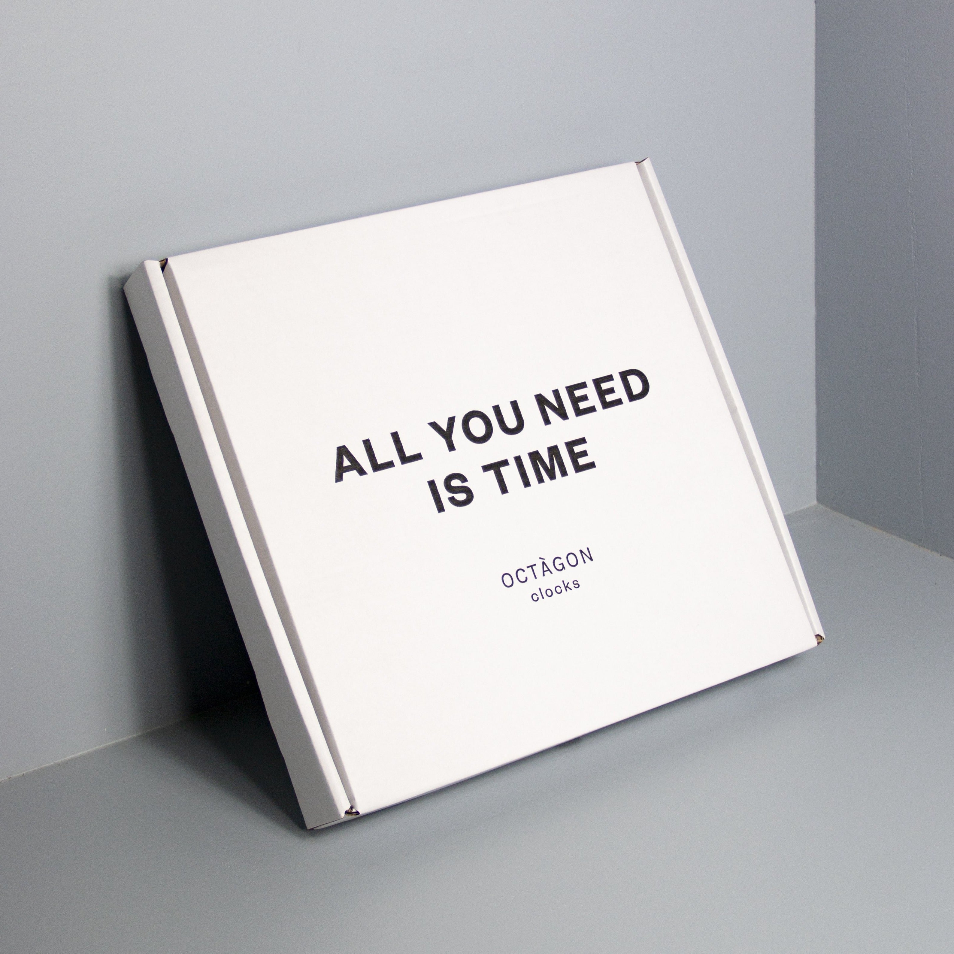 Box with &quot;All you need is time&quot; print leaning on a gray wall | Caja estampada &quot;All ypu need is time&quot; apoyada en una pared gris. | Caixa estampada &quot;All you need is time&quot; recolzada en una paret gris.
