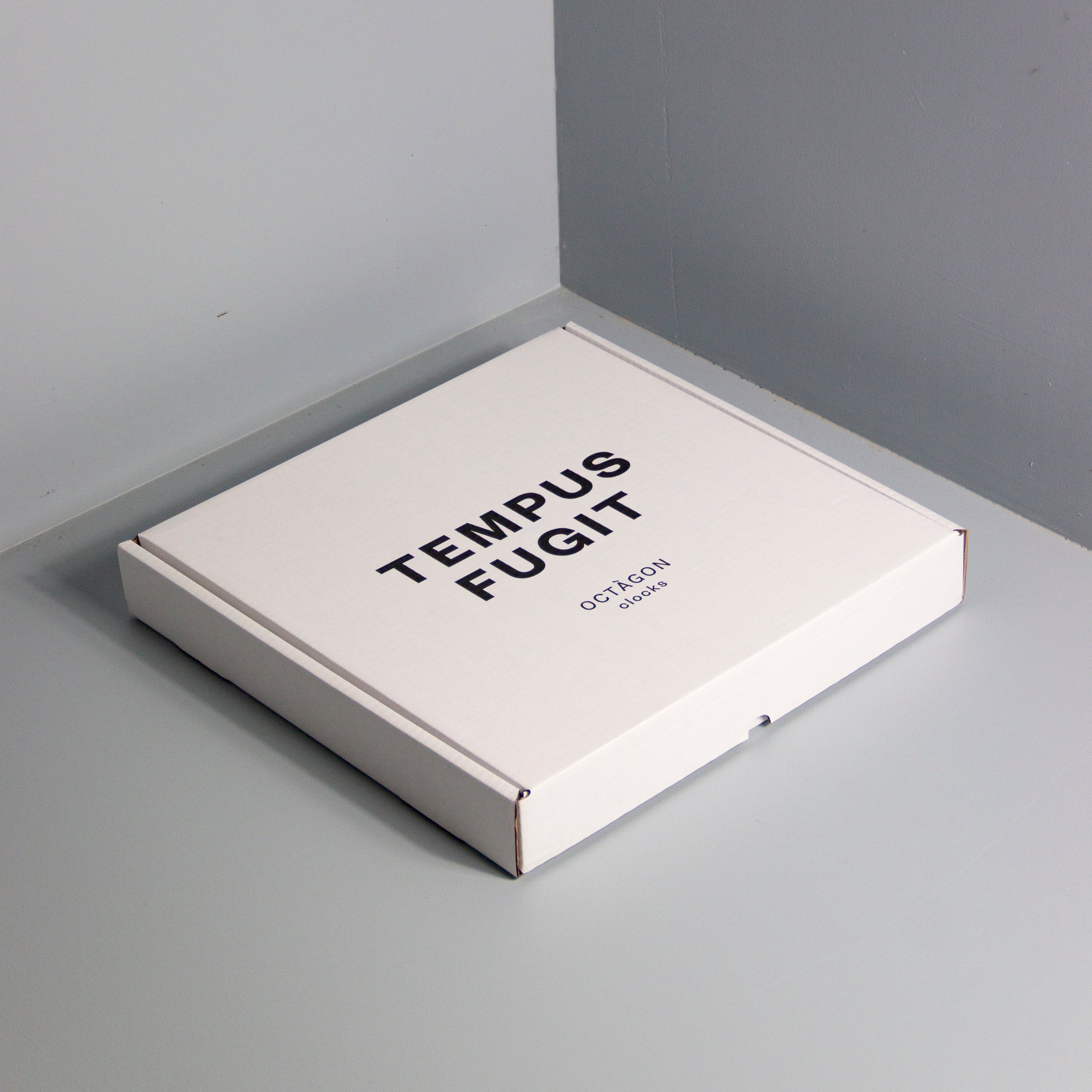 Box with "Tempus fugit" print leaning on a gray wall | Caja estampada "Tempus fugit" apoyada en una pared gris. | Caixa estampada "Tempus fugit" recolzada en una paret gris.
